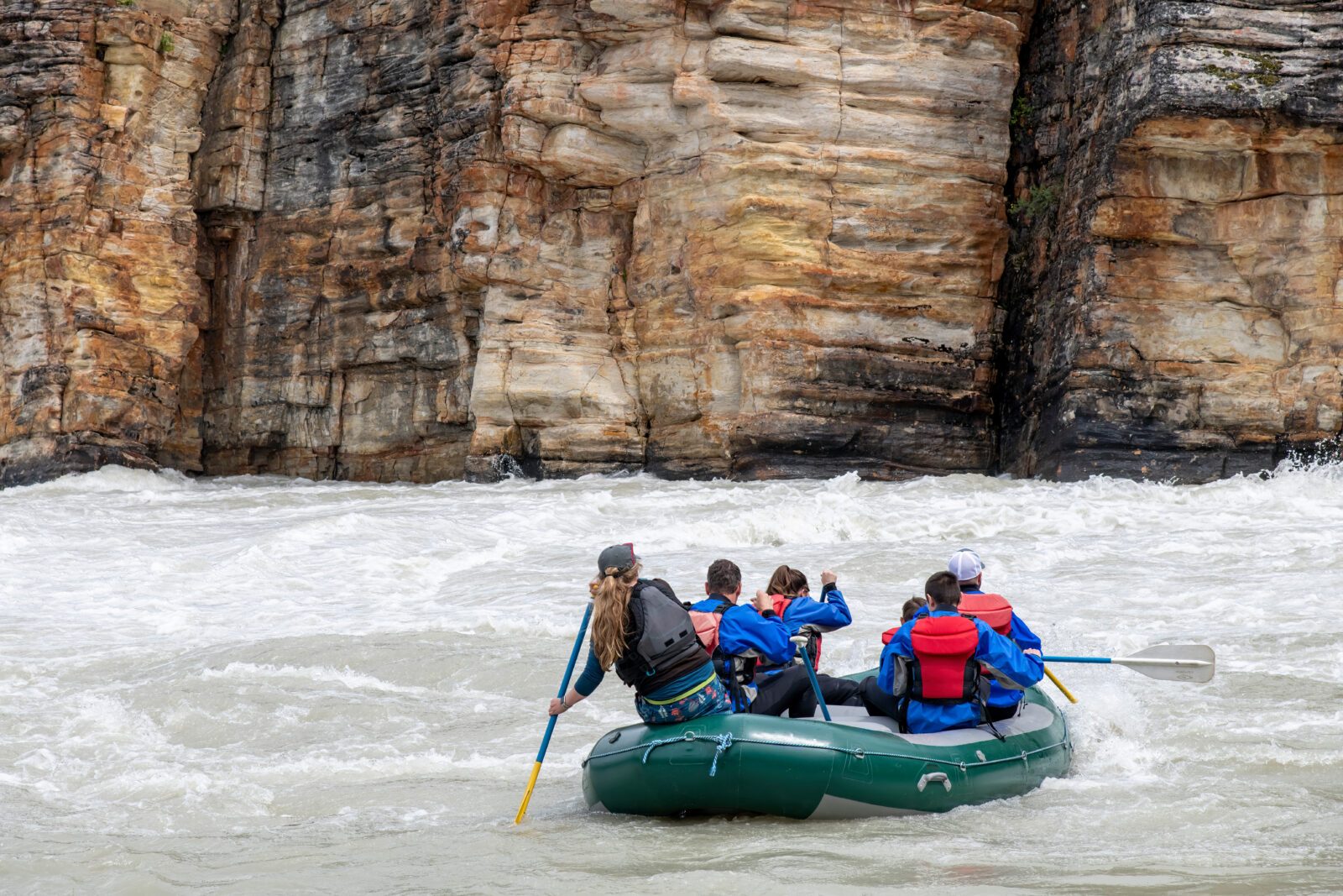 Close up of people in white-water raft on the Athabasca River flowing from nearby Athabasca Falls, Jasper National Park, AB, Canada, with colorful layers of hard quartzite and softer limestone in back